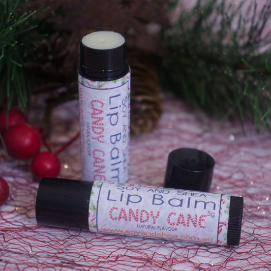 Displayed are two tubes of the "Candy Cane Lip Balm" (Christmas Edition) from Soy and Shea. One tube stands upright with its cap removed, showcasing the nourishing balm inside, and the other is positioned horizontally. Festive decorations with red berries and green pine branches embellish the background.