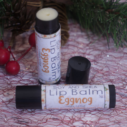 Two tubes of the "Eggnog Lip Balm (Christmas Edition)" from Soy and Shea are beautifully displayed on a decorative surface, surrounded by holiday-themed greenery and red berries. These handmade lip balms are crafted to nourish and protect, with one tube open to reveal the balm inside.