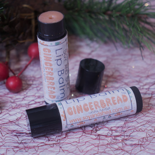 Two tubes of the Gingerbread Lip Balm (Christmas Edition) from Soy and Shea are displayed, with one standing upright and the other lying down, both crowned with black caps. The setting is adorned with festive elements such as pine branches and red berries. Each tube showcases the website "soyandshea.com.au." This lip balm is ideal for keeping your lips nourished during the holiday season.