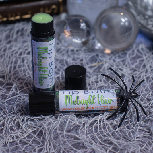 A tube of Soy and Shea's "Midnight Elixir Lip Balm (Halloween Edition)" from www.soyandshea.com.au is displayed with its cap off, lying on a textured gray surface. Nearby, another sealed tube stands upright. Decorative glass orbs and a toy spider surround the tubes, ready to moisturize and nourish lips with natural oils.