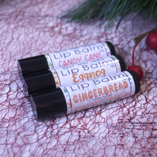 A festive netted surface showcases the Christmas Lip Balm Bundle from Soy and Shea, featuring three limited edition flavors: "Candy Cane," "Eggnog," and "Gingerbread." Greenery and red berries provide a seasonal backdrop for these holiday treats.