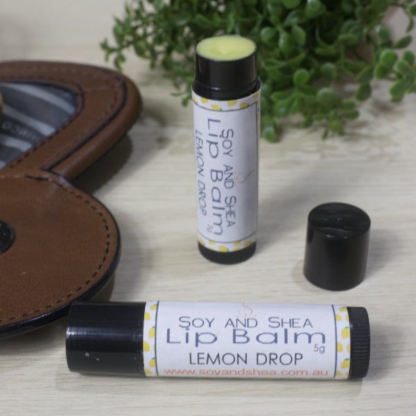 Two black lip balm tubes labeled 'Soy and Shea Lip Balm Lemon Drop' on a wooden surface with a brown leather bag and green plant in the background.  One Lip Balm is lying on its side while the other is standing on its end with the lid removed showing a yellow balm.