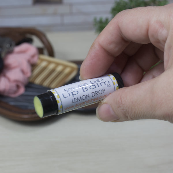 Hand holding a lip balm in a black tube labeled 'Lemon Drop' with a blurred background.  The tube has had its lid removed showing a yellow balm