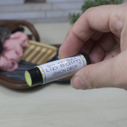 Hand holding a lip balm in a black tube labeled 'Lemon Drop' with a blurred background.  The tube has had its lid removed showing a yellow balm