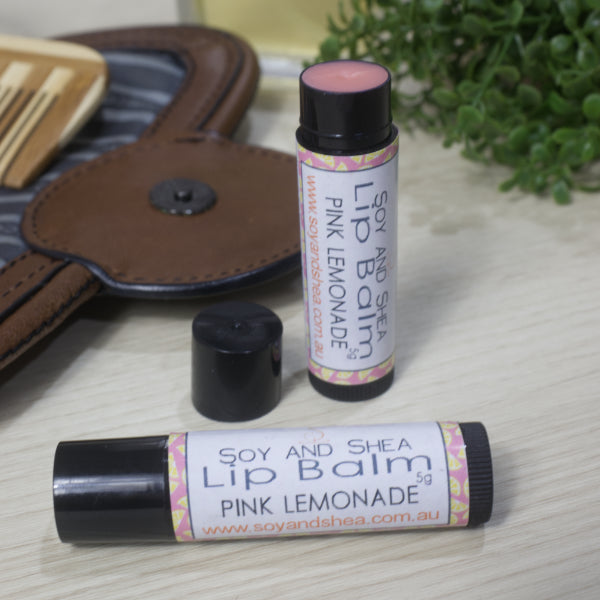Two lip balm tubes labeled 'Soy and Shea' on a surface with a brown leather bag and green plant in the background.  One Lip Balm tube lies on the side while the other stands on its end with the lid placed on the side revealing a pink balm
