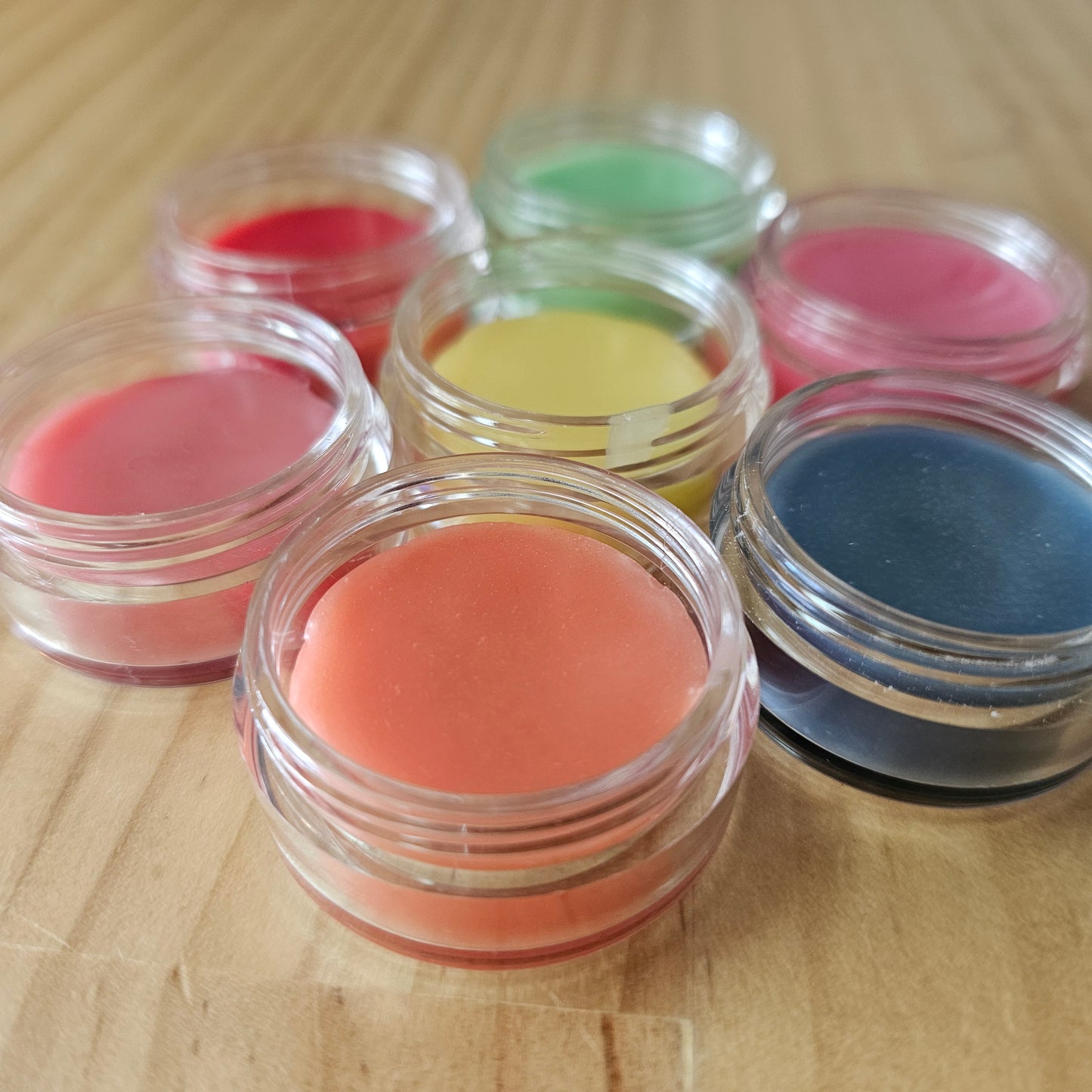 Set of small clear containers filled with colorful lip balm sit on a wooden surface