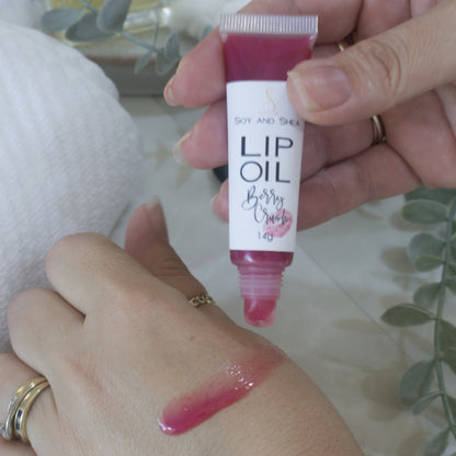 A hand holds a tube of Lip oil next to another hand that has a smear of the oil across the back demonstrating the colour and translucent nature of the oil.  The tube is clear showing a cerise Lip Oil that goes onto the hand with a sheer look.