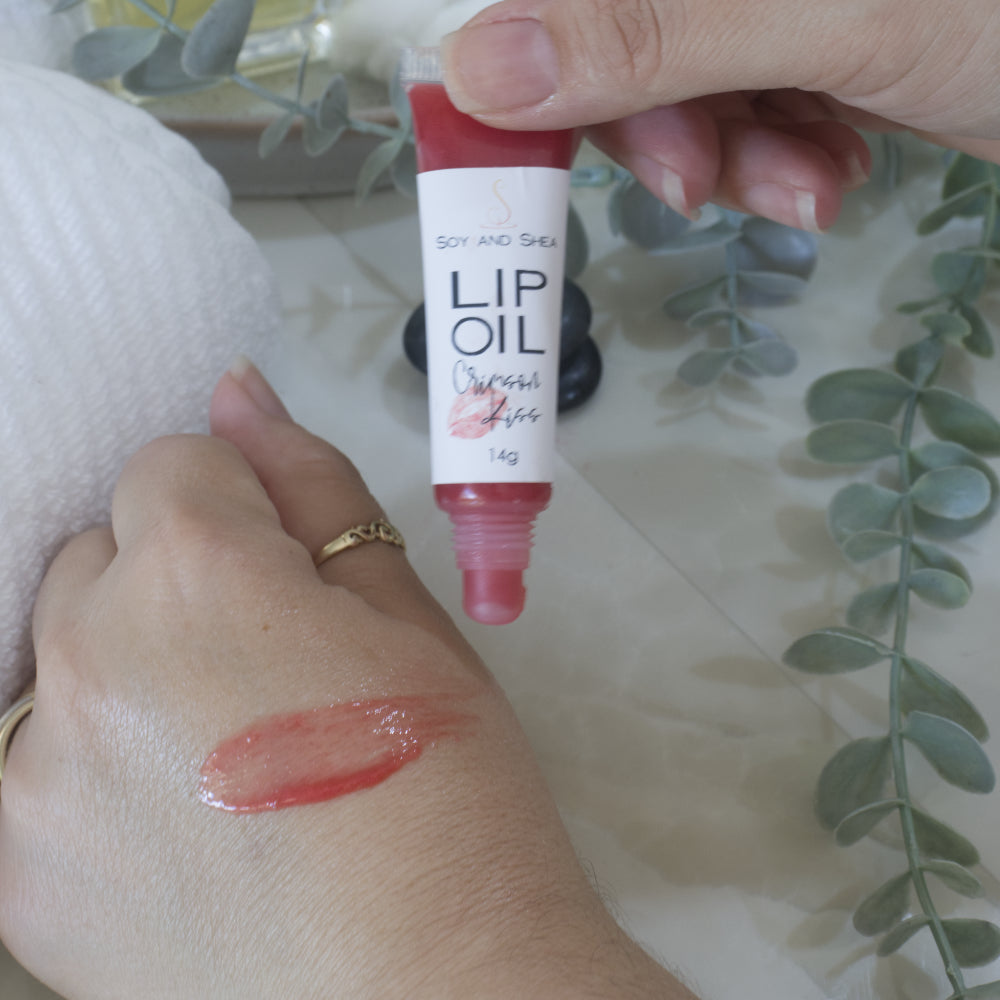 A hand holds a tube of Lip oil next to another hand that has a smear of the oil across the back demonstrating the colour and translucent nature of the oil.  The tube is clear showing a red Lip Oil that goes onto the hand with a sheer look.