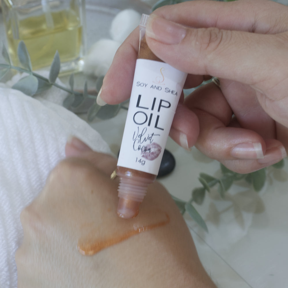 A hand holds a tube of Lip oil next to another hand that has a smear of the oil across the back demonstrating the colour and translucent nature of the oil.  The tube is clear showing a Chocolate Brown Lip Oil that goes onto the hand with a sheer look.