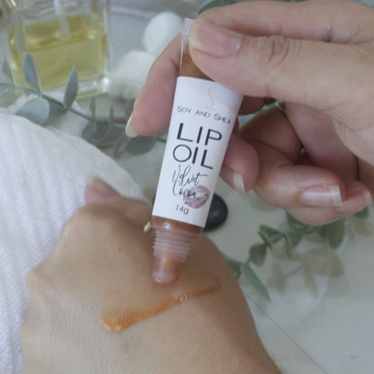 A hand holds a tube of Lip oil next to another hand that has a smear of the oil across the back demonstrating the colour and translucent nature of the oil.  The tube is clear showing a Chocolate Brown Lip Oil that goes onto the hand with a sheer look.