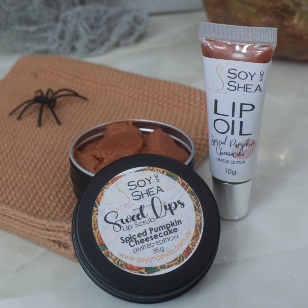 Soy and Shea lip care products arranged on a light surface: a black tin of spiced pumpkin cheesecake lip scrub with the lid removed and resting on the side of the tin revealing a bronze scrub inside.  To the right stands a tube of spiced pumpkin lip oil. A decorative spider and burlap props are present.