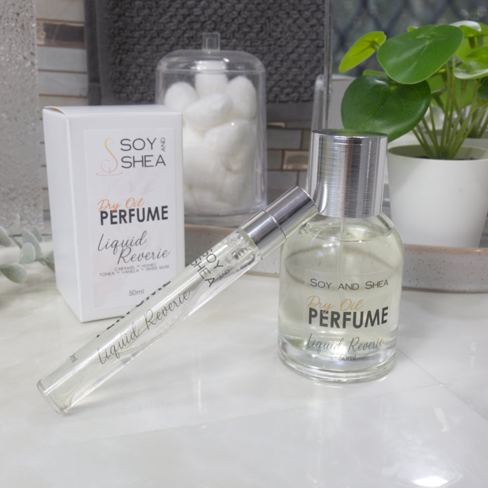 A selection of perfume bottles are presented on a bathroom countertop.  A clear, narrow, tall bottle labeled as 10ml with a silver cap stands rests at an angle against a clear round, short bottle with silver lid labelled as 50ml.  Behind sits a white box with label identifying as Dry Oil Perfume Liquid Reverie.