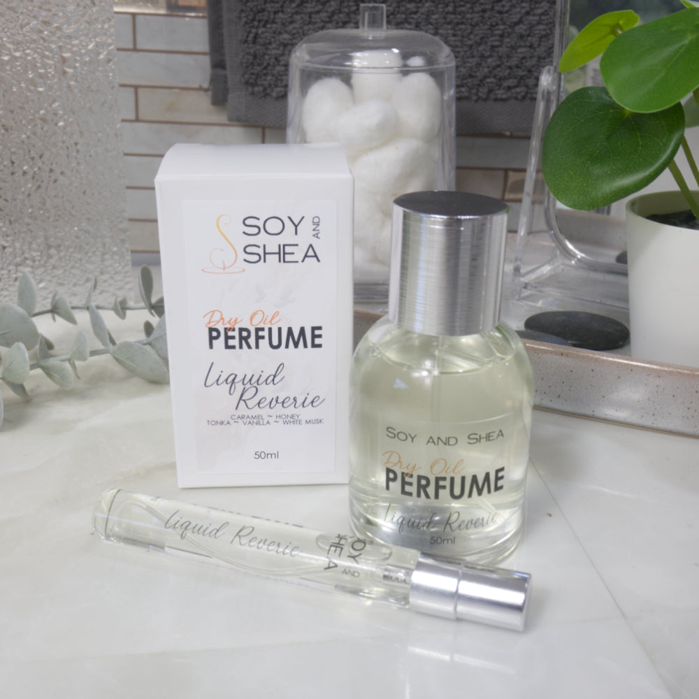 A selection of perfume bottles are presented on a bathroom countertop.  To the front is a clear, narrow, tall bottle labeled as 10ml with a silver cap and behind is a clear round, short bottle with silver lid labelled as 50ml.  Behind sits a white box with label identifying as Dry Oil Perfume Liquid Reverie.