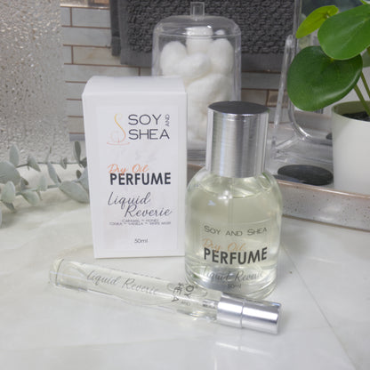 A selection of perfume bottles are presented on a bathroom countertop.  To the front is a clear, narrow, tall bottle labeled as 10ml with a silver cap and behind is a clear round, short bottle with silver lid labelled as 50ml.  Behind sits a white box with label identifying as Dry Oil Perfume Liquid Reverie.