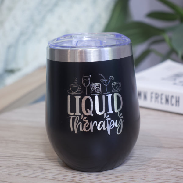 A black metal drink tumbler with silver rim and engraved with LIQUID THERAPY and a collection of various drinks sits on a wooden table with book against a blurred background with greenery.