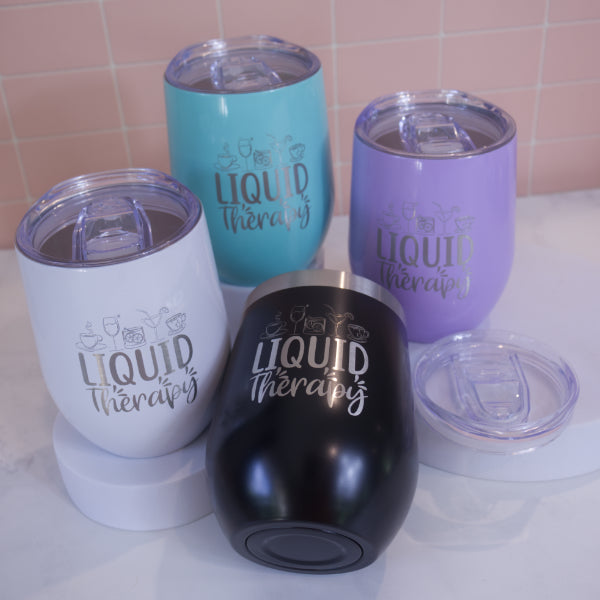 A collection of insulated metal tumblers in various colors, each engraved with the phrase 'Liquid Therapy' on the side.  Each tumber has a clear lid with sliding cover over the mouth piece