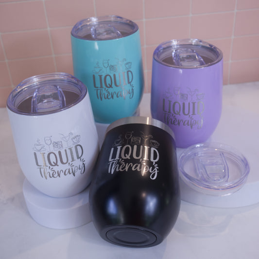 A collection of insulated metal tumblers in various colors, each engraved with the phrase 'Liquid Therapy' on the side.  Each tumber has a clear lid with sliding cover over the mouth piece