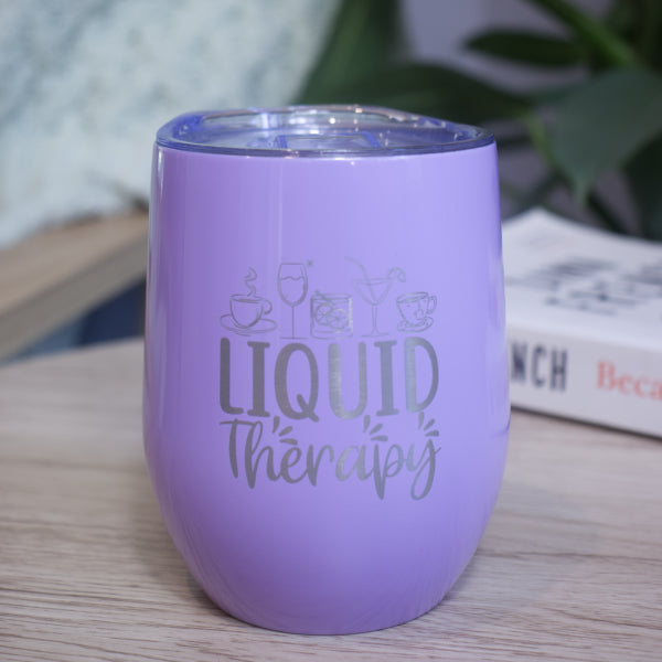 A lilac metal drink tumbler engraved with LIQUID THERAPY and a collection of various drinks sits on a wooden table with book against a blurred background with greenery.
