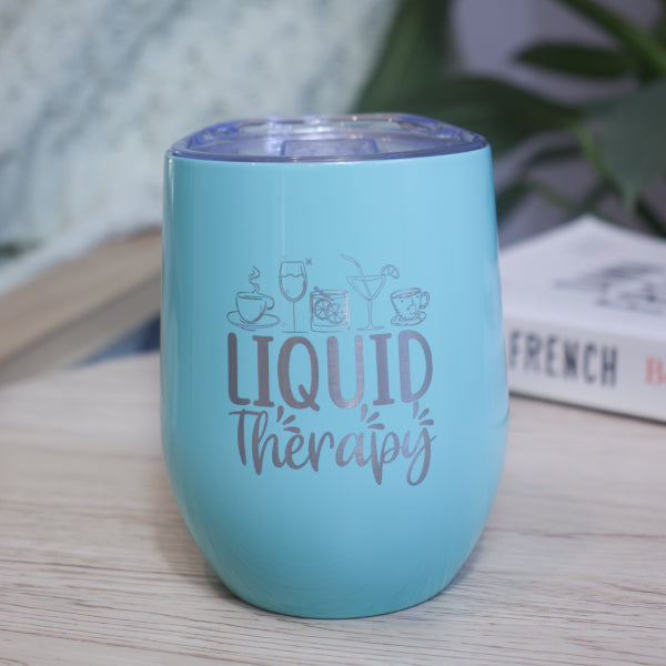 A teal metal drink tumbler engraved with LIQUID THERAPY and a collection of various drinks sits on a wooden table with book against a blurred background with greenery.