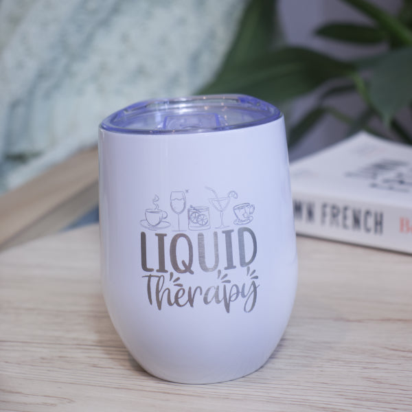 A white metal drink tumbler engraved with LIQUID THERAPY and a collection of various drinks sits on a wooden table with book against a blurred background with greenery.