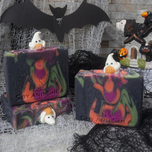 Three bars of soap sit on a bench surrounded by halloween props including a haunted house and bat.  One bar of soap stands upright to the front right, while to the left, another soaps lays flat with a third standing upright on it.   The soaps have a black base colour with drops of neon orange, purple and black colour.  Each soap has a small ghost embellishment holding onto a pumpkin