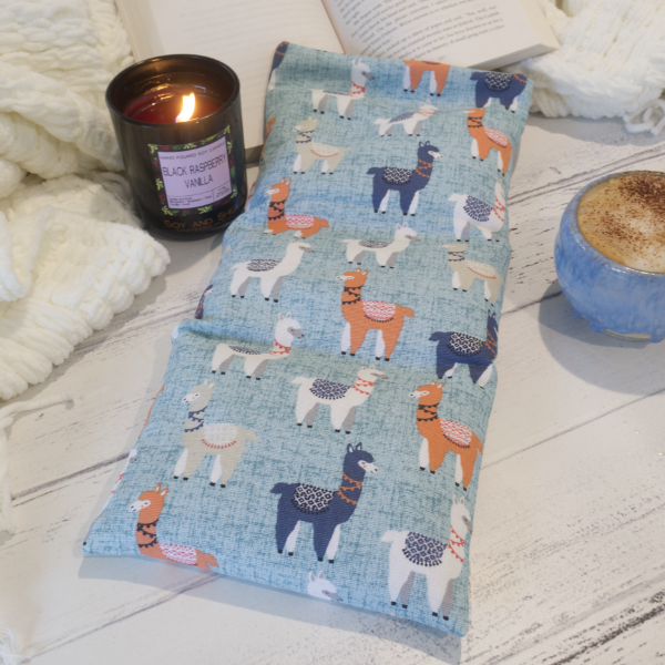 A long rectangular heat pack with a blue fabric cover, decorated with a pattern of multicolored llamas, placed on a wooden surface alongside a candle and a cup of coffee.