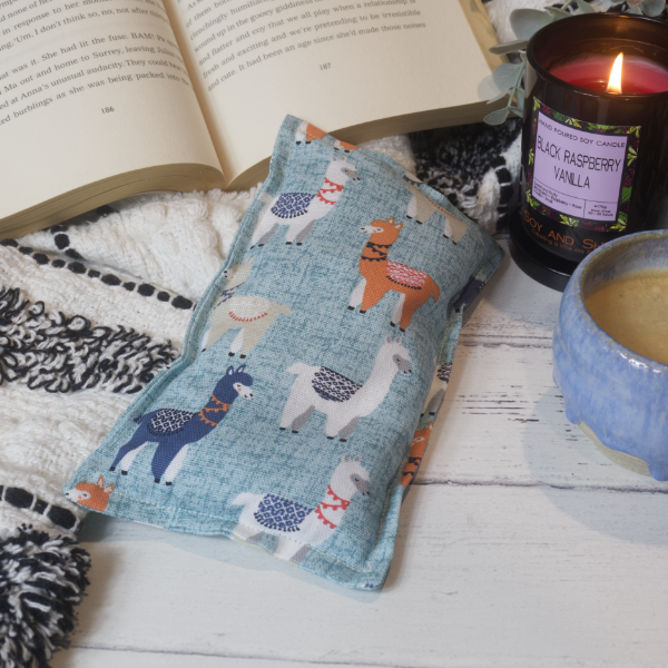 A mini rectangular heat pack rests against a black and white blanket and open book.  Next to it is a burning candle and filled coffee cup evoking a comfy scene.   The Heat Pack has a pale blue background with teal speckles along with illustrations of orange, blue or white llamas with blankets on their backs