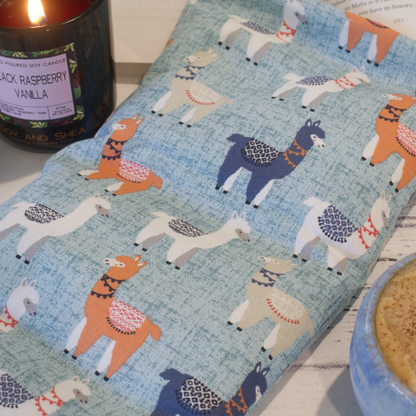 A close up picture of the Llama Drama Heat pack sitting next to a burning candle.   The Heat Pack has a light blue speckled background and features drawings of llamas in blue, orange, white and grey