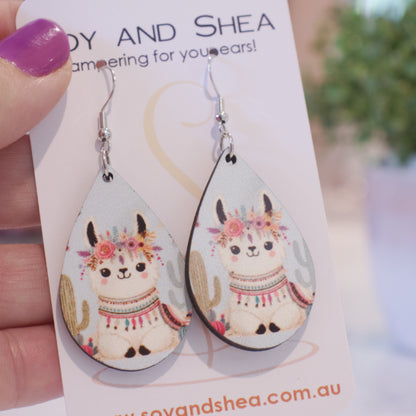 A hand holds a white card branded with SOY AND SHEA with a pair of earrings hanging on hooks.  The earrings are teardrop shaped featuring an illustration of a llama lying down surround by cactus.  The llama has a garland of flowers around its head and blanket on back with decorations around the neck giving it a boho appearance.