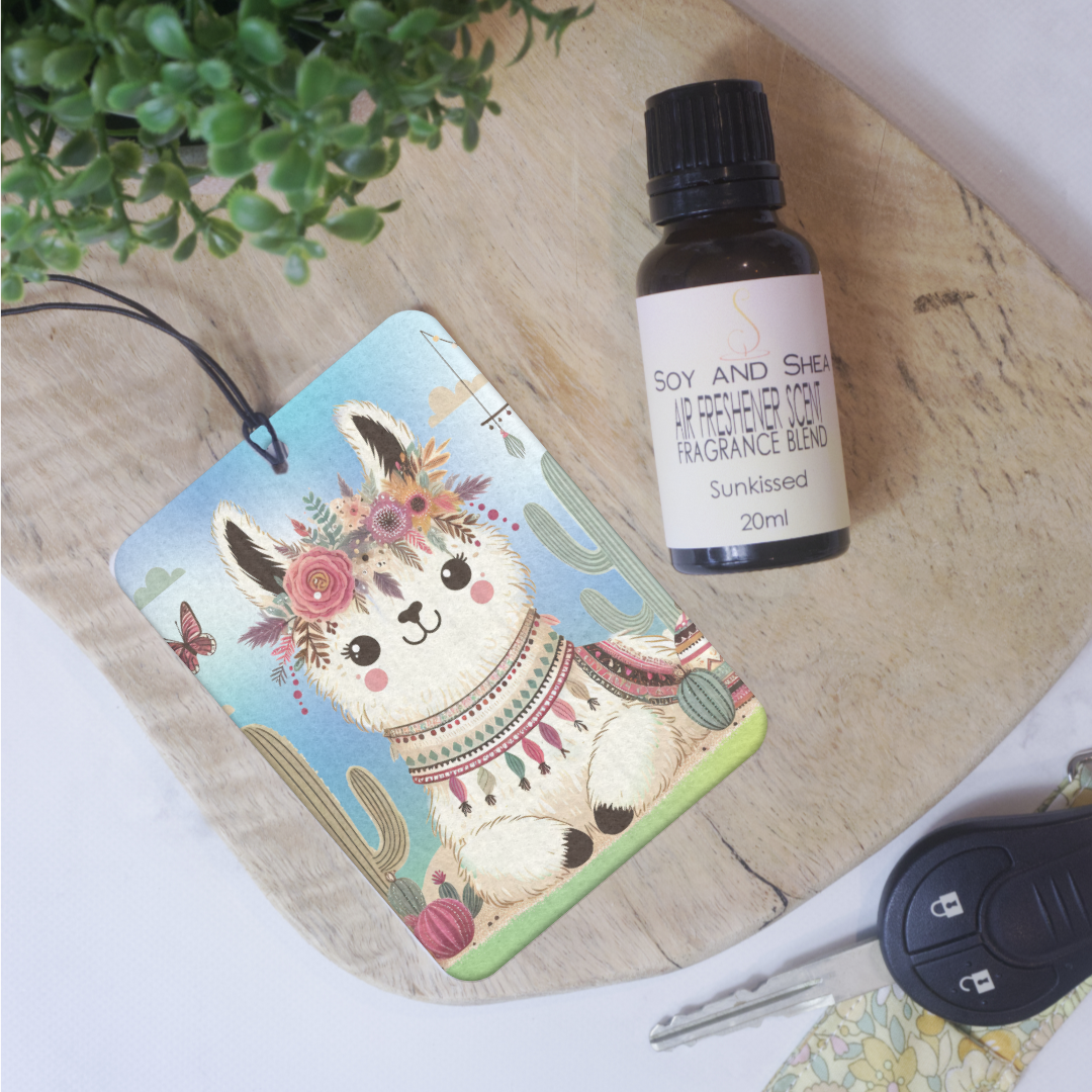 A car air freshener featuring an illustration of a boho-style llama with flowers and a colorful patterned scarf lies on a wooden board next to a bottle of air freshener scent and a car key