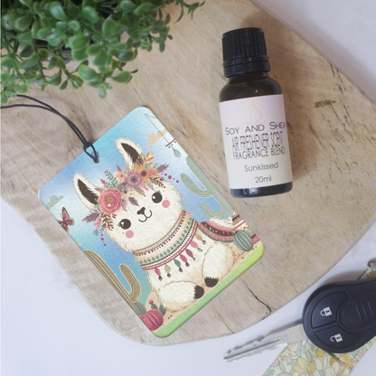 A car air freshener featuring an illustration of a boho-style llama with flowers and a colorful patterned scarf lies on a wooden board next to a bottle of air freshener scent and a car key