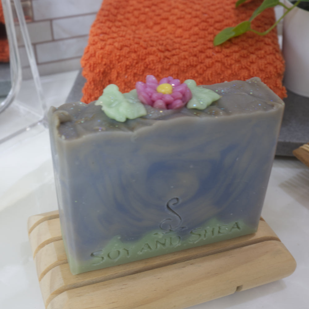 A close up image of the Lotus Flower Soap bar sitting on a wooden soap dish with towel in the background.  The focus is a pink flower with yellow center and surrounded by leaves resting on the top of the soap, seemingly made of soap.   The main colour of the soap is a swirling pattern of various blue shades with a think uneven, green base.