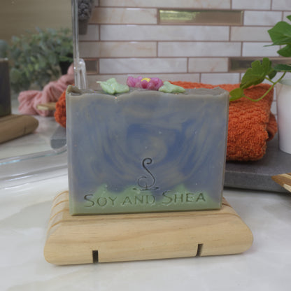 A bar of soap stands on a wooden soap dish on a bathroom countertop surrounded by a towel, mirror and greenery.   The soap features a thin, uneven green layer across the base with the top beings various blue shades swirled together.  The top of the soap has a soap embellishment of a pink flower and leaves.
