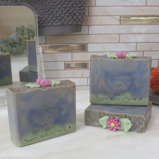 A collection of three soap bars are displayed on a marble surface with a tiled wall background with towel and mirror.  One soaps lies flat displaying the top of the soap which features a soap embellishment of a pink flower with yellow center and green leaves.  Another soap stands upright on this bar with a third bar placed to the left.  The soaps features a thin, uneven green layer across the base with the top beings various blue shades swirled together.  