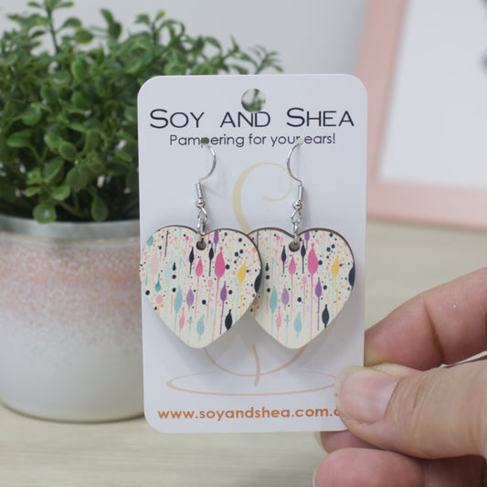 Heart-shaped earrings with a colourful paint splatter design on a 'Soy and Shea' branded card with a plant in the background.