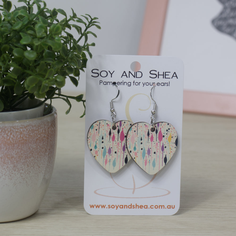 Heart-shaped earrings with a colourful paint splater design on a Soy and Shea branded card with a plant in the background