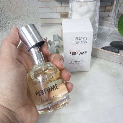 A hand holds a bottle of Soy Shea Dry Oil Perfume Lumina with a silver spray cap pulled up to reveal a silver atomiser.  Behind is its white packaging box on a bathroom counter.