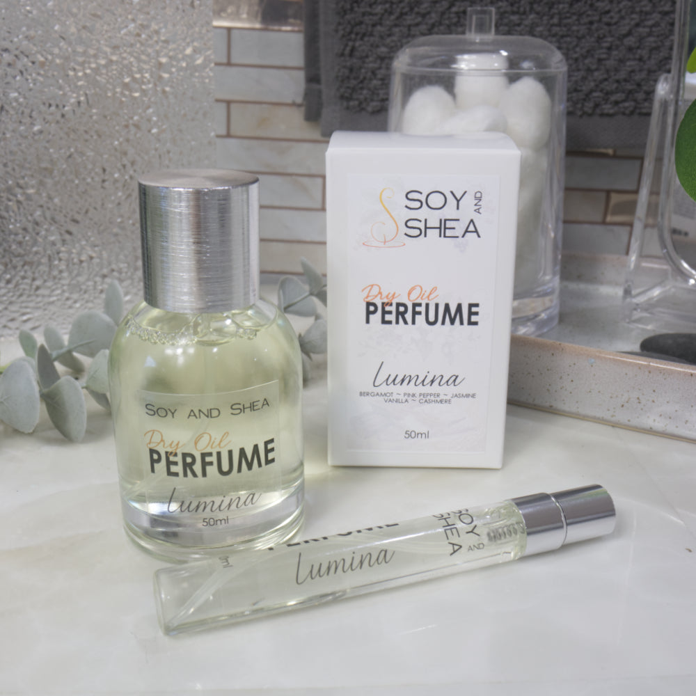 A selection of perfume bottles are presented on a bathroom countertop.  To the front is a clear, narrow, tall bottle labeled as 10ml with a silver cap and behind is a clear round, short bottle with silver lid labelled as 50ml.  Behind sits a white box with label identifying as Dry Oil Perfume Lumina.