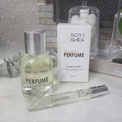 A selection of perfume bottles are presented on a bathroom countertop.  To the front is a clear, narrow, tall bottle labeled as 10ml with a silver cap and behind is a clear round, short bottle with silver lid labelled as 50ml.  Behind sits a white box with label identifying as Dry Oil Perfume Lumina.