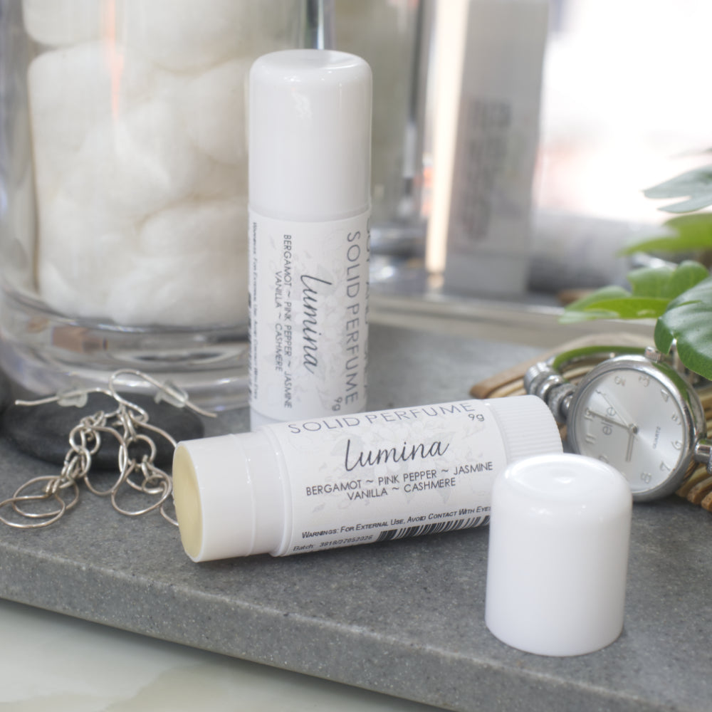 Two white tubes of Solid Perfume are displayed on a grey stone tray along with silver earrings, watch and greenery.   One tube lays flat displaying the fragrance notes while another tube stands upright behind it. The flat tube has the lid removed and placed to the front right revealing a beige balm inside.