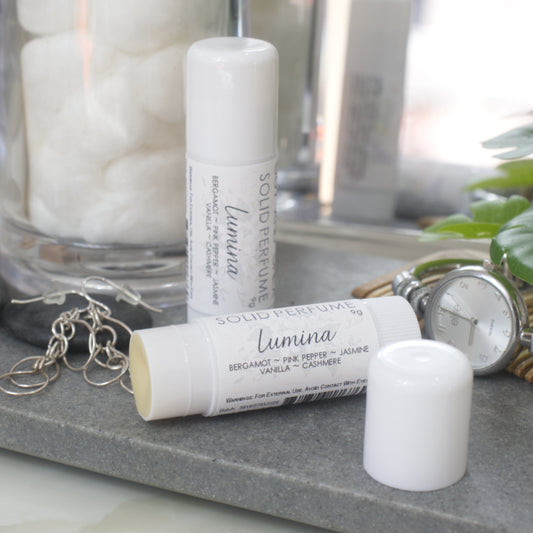 Two white tubes of Solid Perfume are displayed on a grey stone tray along with silver earrings, watch and greenery.   One tube lays flat displaying the fragrance notes while another tube stands upright behind it. The flat tube has the lid removed and placed to the front right revealing a beige balm inside.