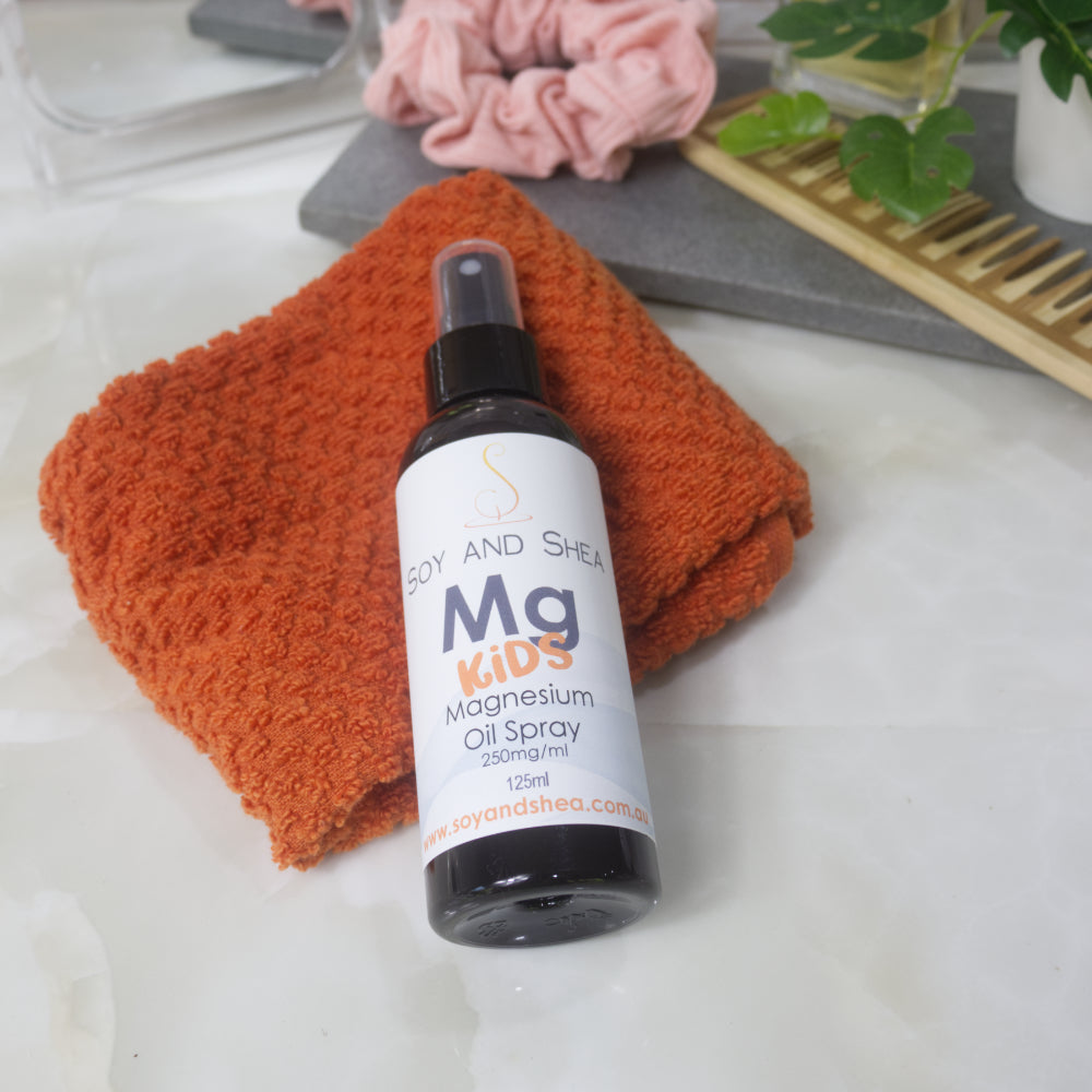 A bottle of Kid Magnesium Oil Spray lays on resting on an orange cloth on a marble bench top with bathroom accessories blurred in the background.  The bottle is amber and features an atomiser spray lid.