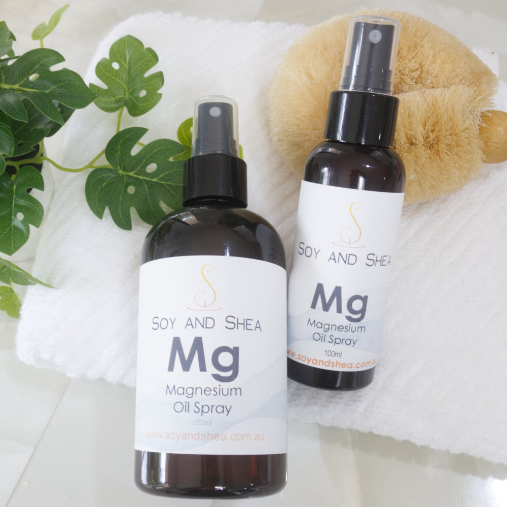 A large and small bottle of Soy and Shea Magnesium Oil Spray on a white towel with a plant in the background.  The bottles are amber with a white label and spray nozzle