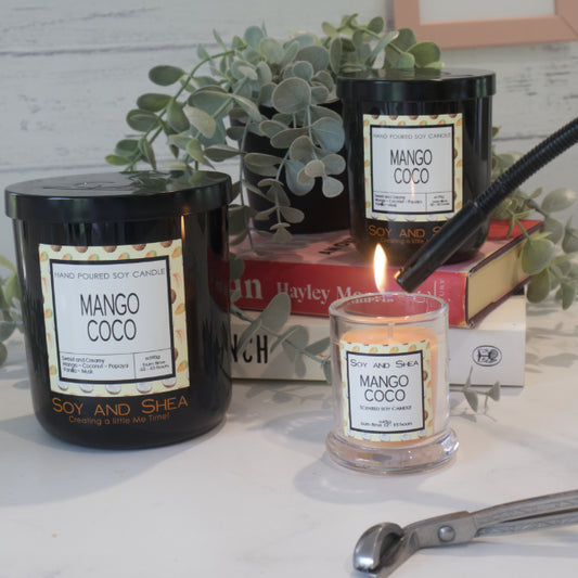 Three Soy and Shea branded candles all with labels reading 'Mango Coco' sit on a wooden surface.  A small candle with clear glass and orange wax sits in front of a stack books, with a medium sized black jar sitting upon them.  The third black jar is the largest and sits to the left.  The small candle is has a lighter holding a flame towards the wick.