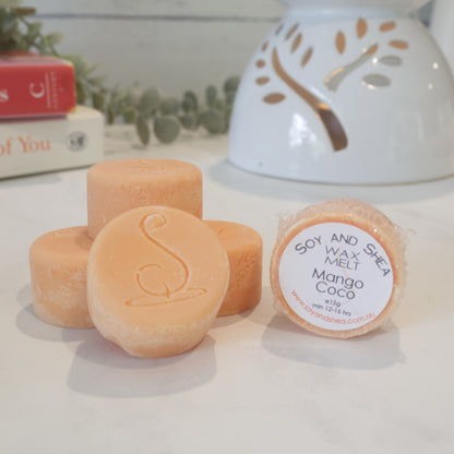 Three cylindrical orange Melt Pucks are stacked up each other with the soy and shea logomark stamped on the tops.  To the right is a fourth melt puck that rests on its side with a label identifying it as Mango Coco.  Behind is a stack of books next to a lit oil warmer.