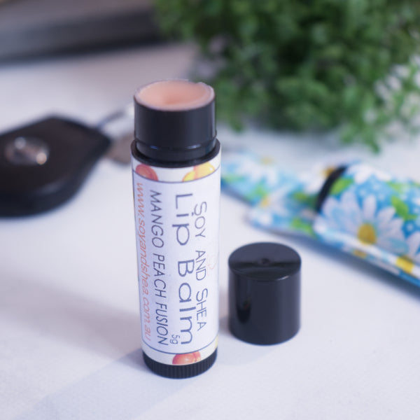 A black tube with colourful label reading LIP BALM MANGO PEACH FUSION stands upright on a white counter with a blurred background.  The black cap has been removed and sits to the side revealing a peach coloured balm