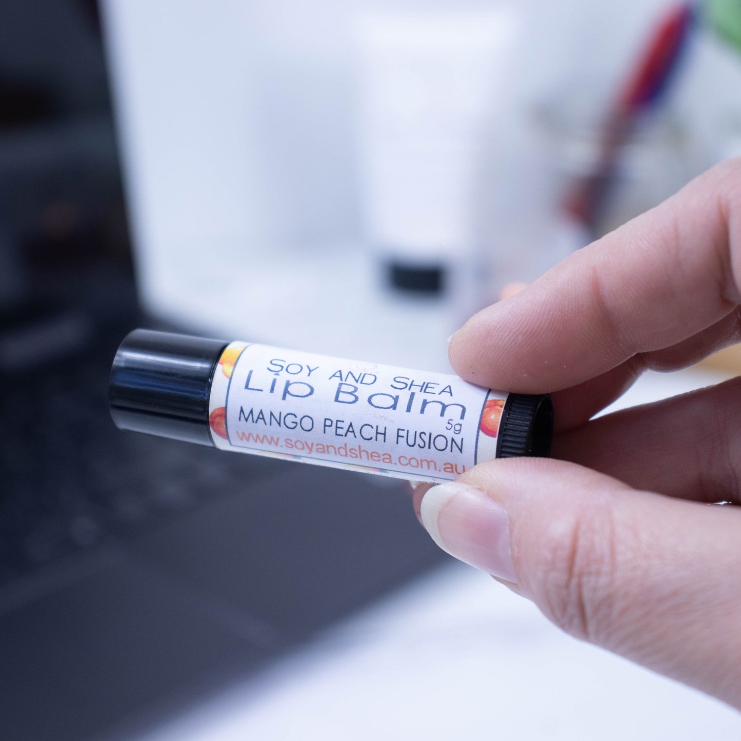 A hand holds a black tube of lip balm against a blurred background with an open laptop computer on a white surface.  The label has text LIP BALM MANGO PEACH FUSION on white background with a border featuring watercolour image of peach and mango.