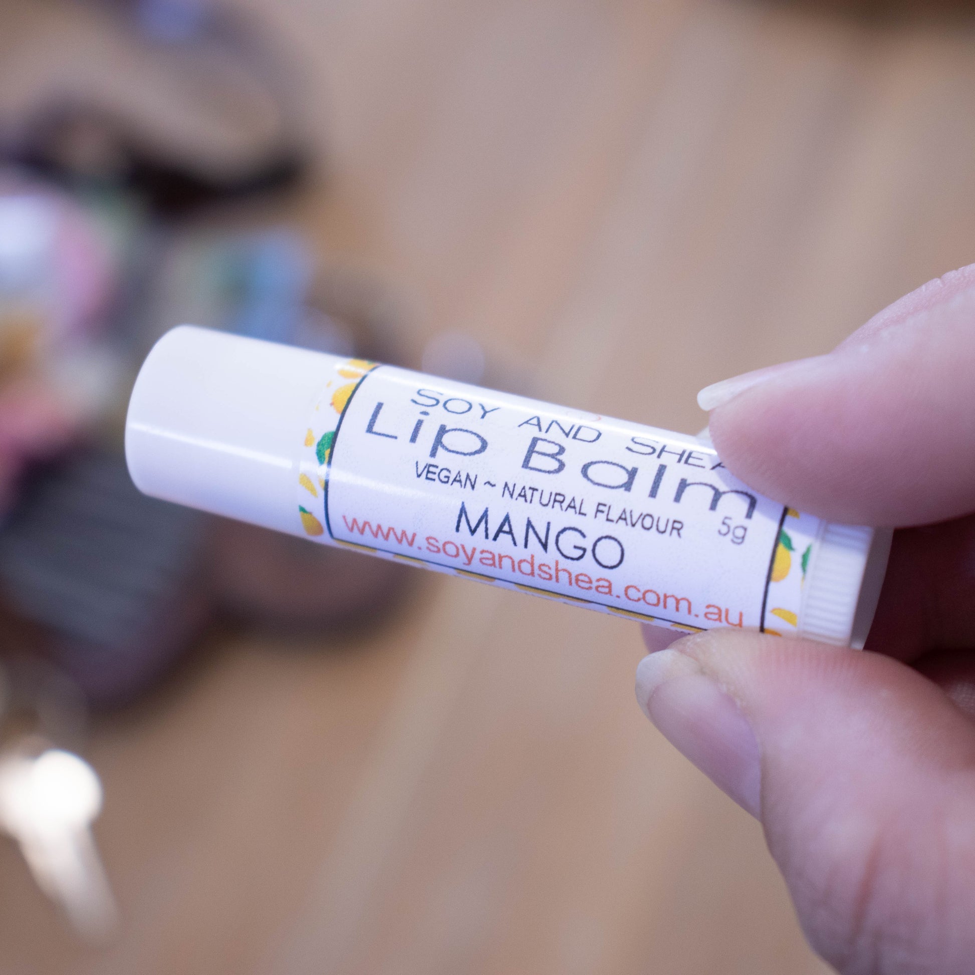 A hand holds a white tube labelled lip balm over a blurred image of an open handbag with contents spilling onto the table.  The lip balms features a white label with illustrations of small mangoes