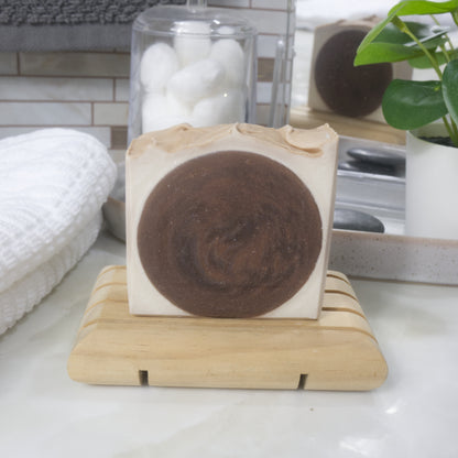 A rectangular bar of soap with a dark brown circular swirl in the center and light beige edges, displayed on a small wooden soap dish on a bathroom counter. A white towel, glass jar, and potted plant are visible in the background.