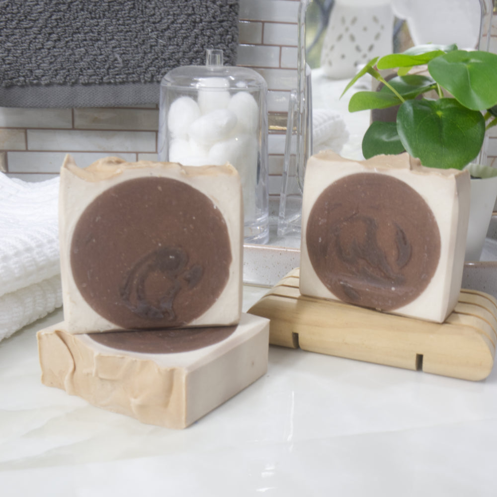 A collection of 3 soaps are displayed on a marble bathroom counter along with a towel, plant and jar of cotton balls.  One soap lays flat on the bench with another standing upright on it.  To the back left is a third soap standing on a wooden soap dish.  The soaps have a dark brown circle in the center with a cream surround of soap